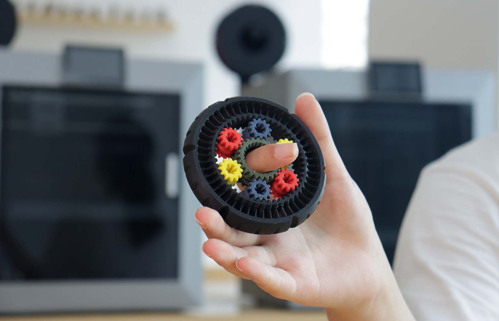 Hand holding a 3D printed planetary gear ring with colorful spur gears, desktop 3D printers blurred in the background