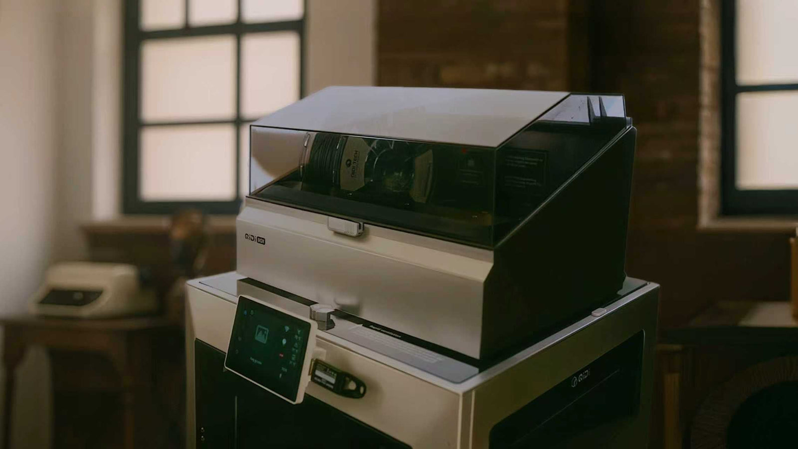 Close‑up of an enclosed desktop 3D printer with a flip‑up top cover and touchscreen, designed for safe, low‑noise printing at home or office