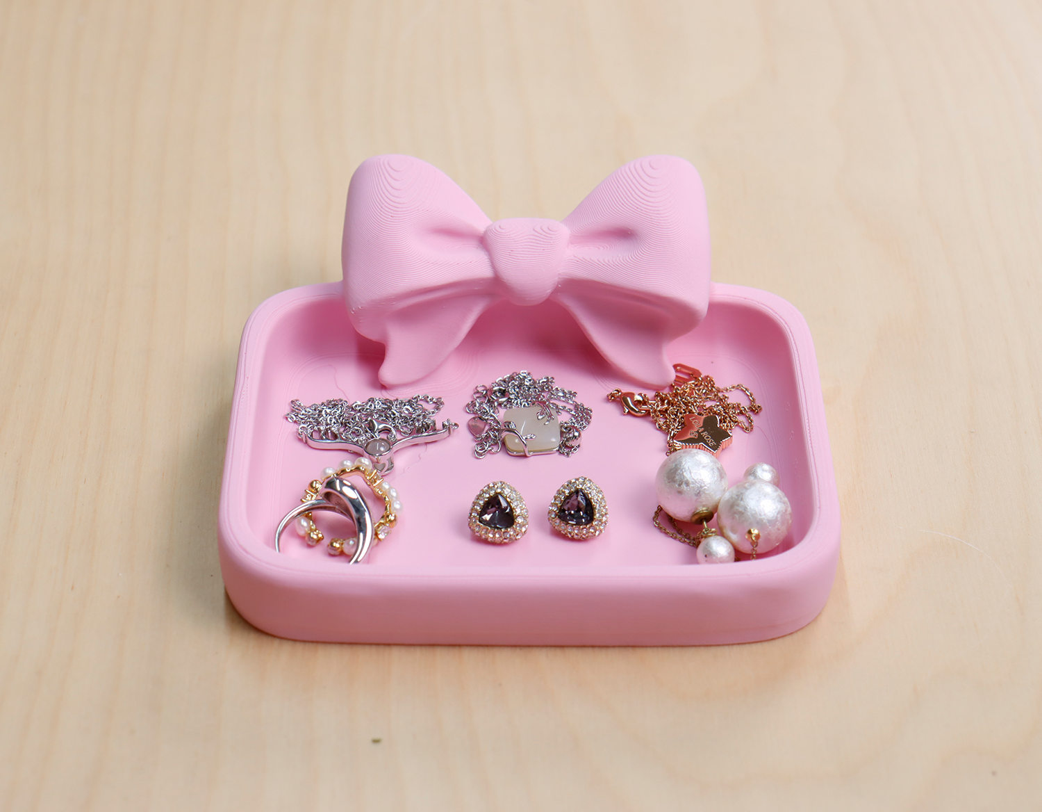 3D‑printed pink bow jewelry tray neatly displaying rings, earrings, and pearls on a wooden tabletop