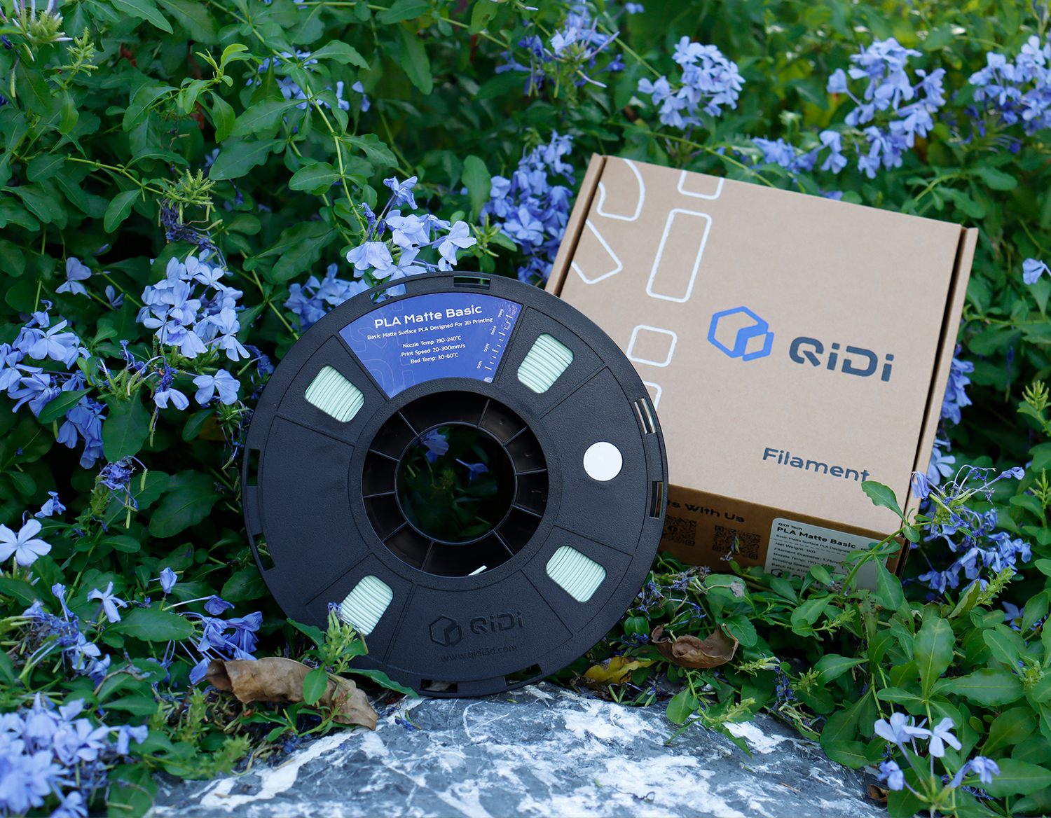 QIDI PLA  3D printer filament spool with branded box, displayed outdoors among purple flowers.